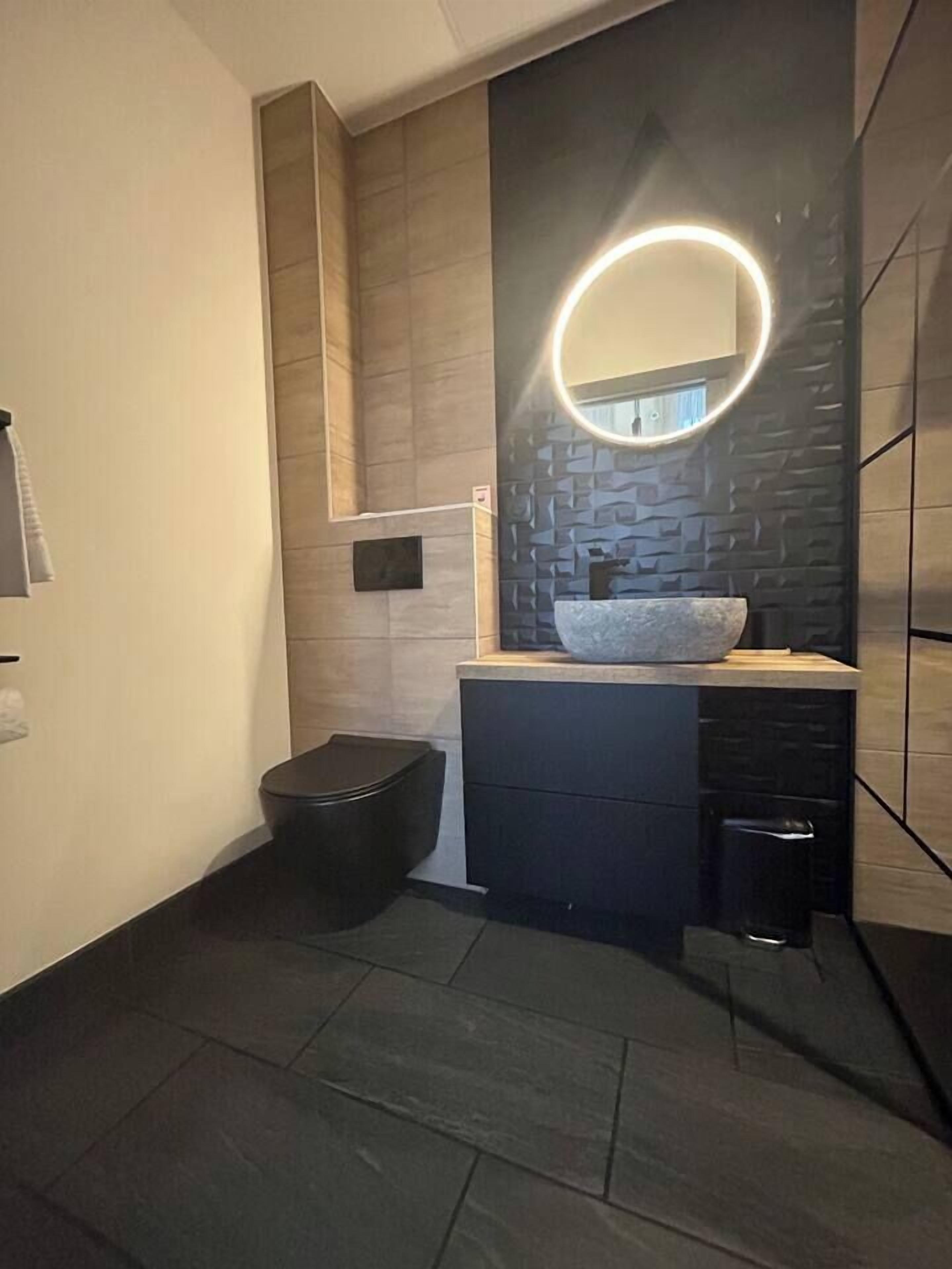 A modern bathroom with a stone sink, a black trash bin, and a wall-mounted mirror with a circular light.