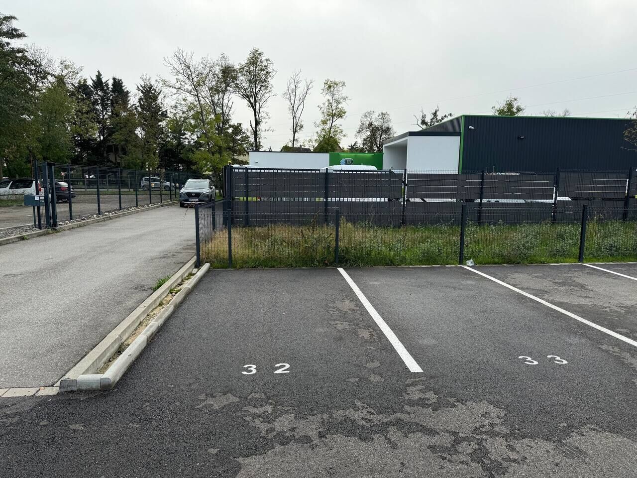 Un parking avec des places délimitées, une clôture et un bâtiment en arrière-plan.
