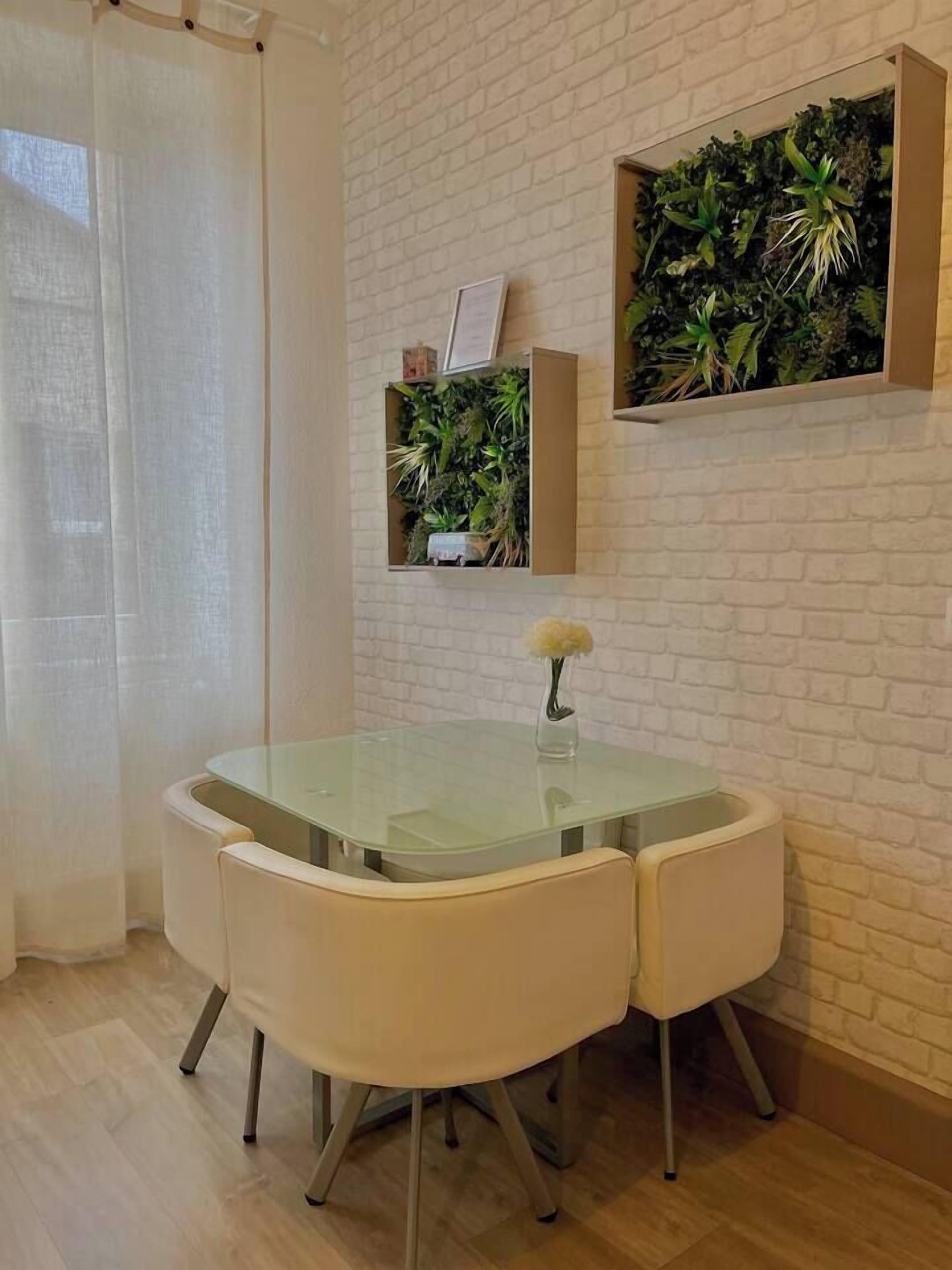 Un coin repas comprenant une table avec un plateau en verre, deux chaises, une petite plante en pot et une étagère murale agrémentée de plantes vertes.