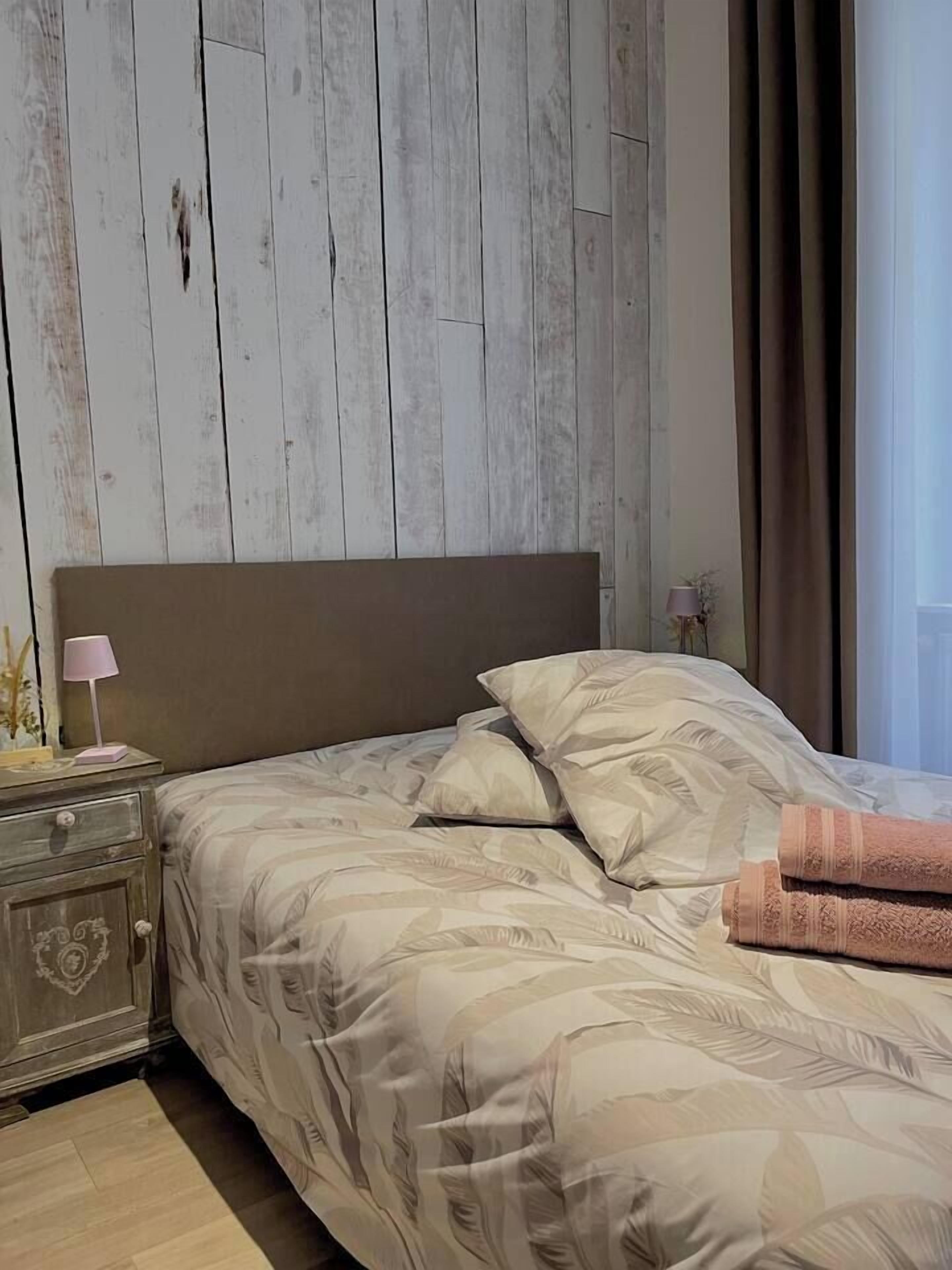 A bedroom with a wooden headboard, a bed with patterned bedding, a nightstand with a lamp, and a window with curtains.