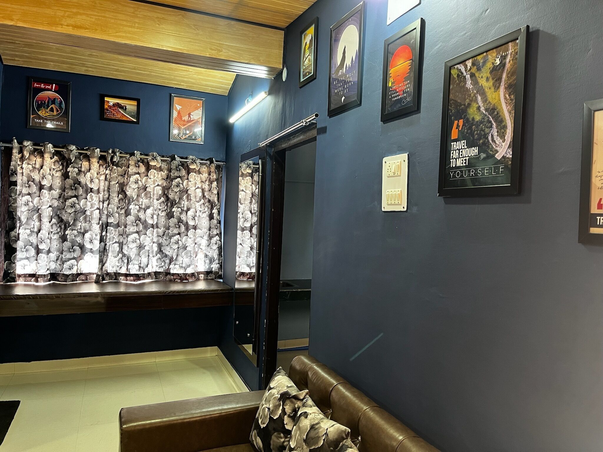 A room with a dark blue wall, a wooden ceiling, and a floral curtain. There are several framed posters on the wall, a brown leather bench, and a light switch.