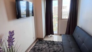 A modern living room with a black sofa, a flat-screen TV mounted on the wall, a potted plant, and a large window with curtains. - Unistay Coast Res R31 with Bal-Sunset Vw (Pasay)