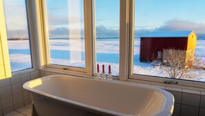 A bathroom with a bathtub, a window, and a view of a snowy landscape and a building. - Villa Lofoten (Bøstad)