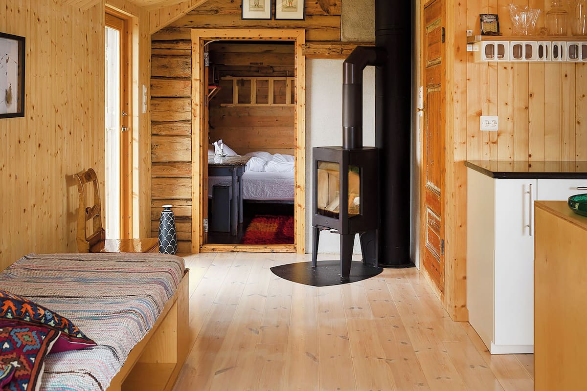 Interior de una cabaña de troncos con cama, chimenea, zona de cocina y puerta que da a otra habitación.