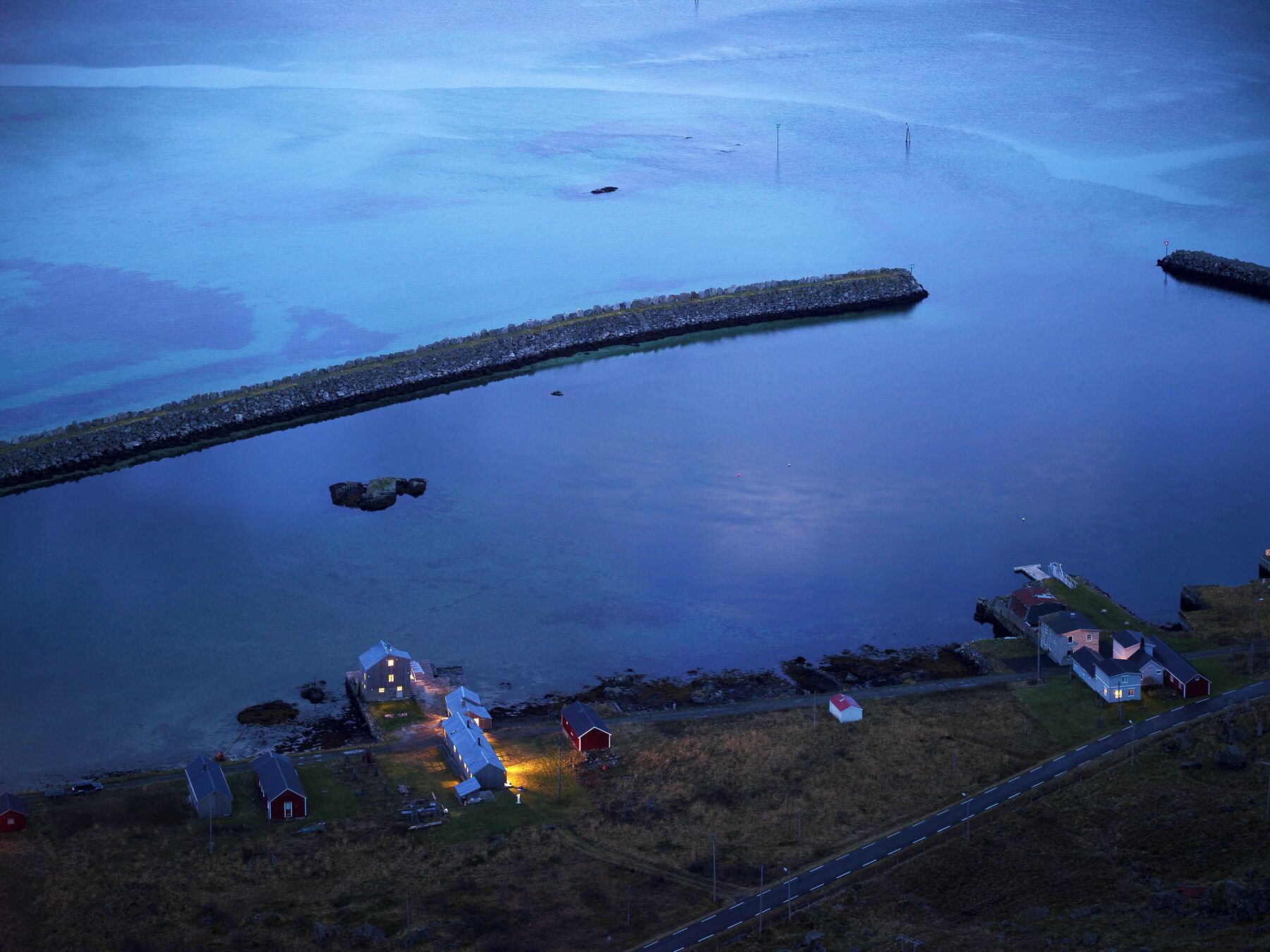 Una zona costera con un muelle que se adentra en aguas tranquilas, rodeada de casas y una carretera.