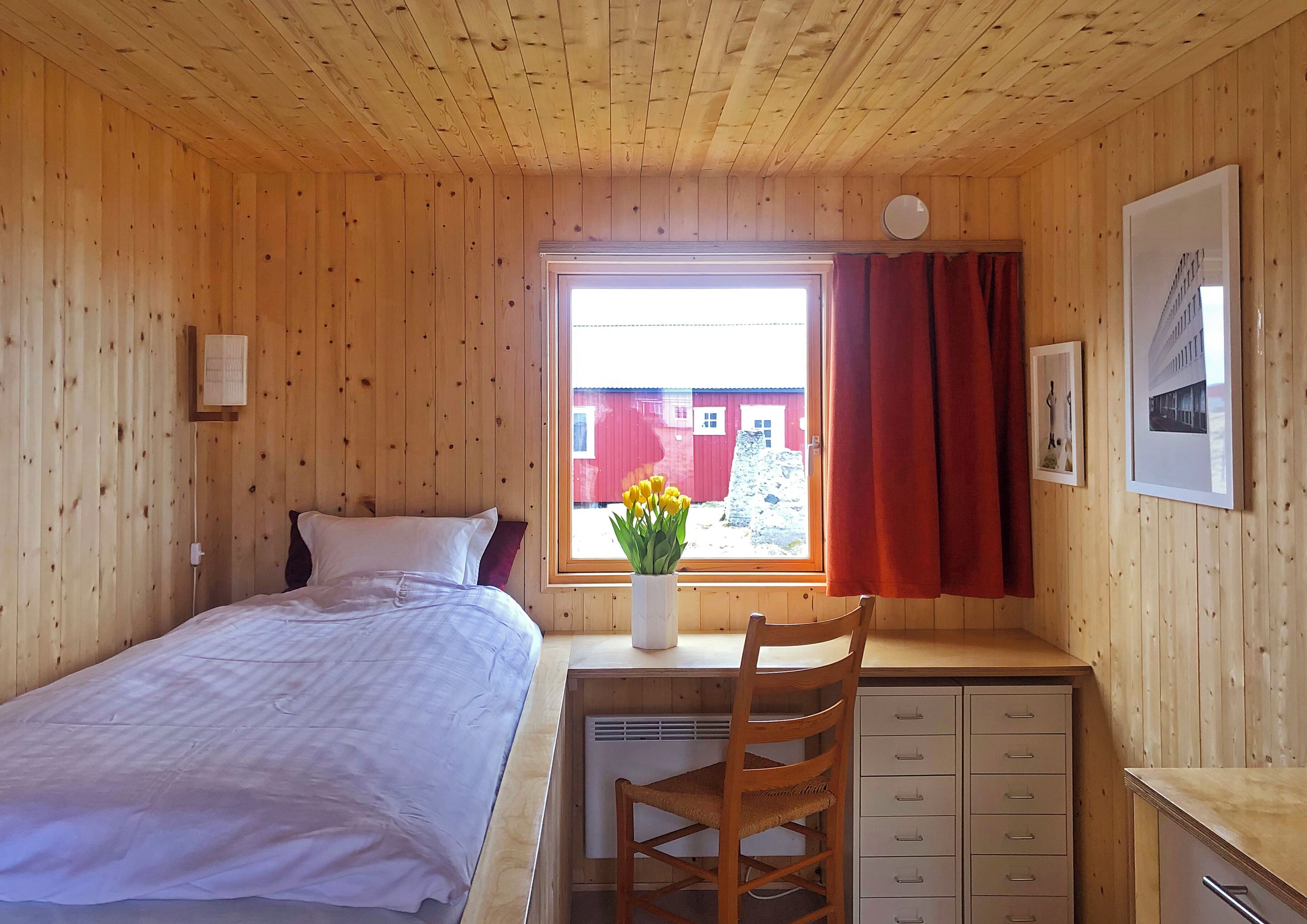 Ett rum med träpanel, en säng, ett skrivbord, en stol, ett fönster med gardiner och en krukväxt.