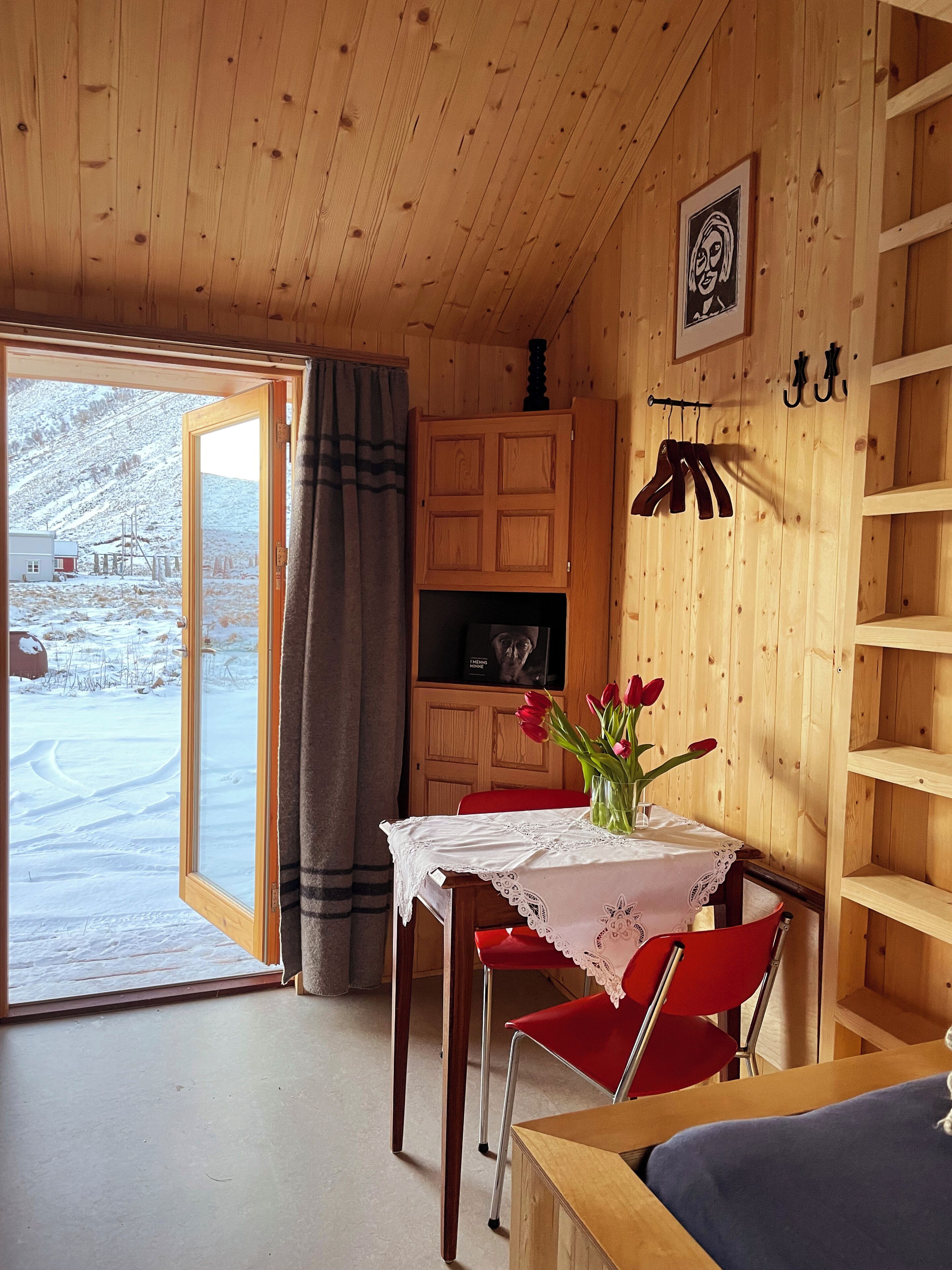 A wooden interior with a table, chairs, a TV, and a view of a snowy landscape through an open door.