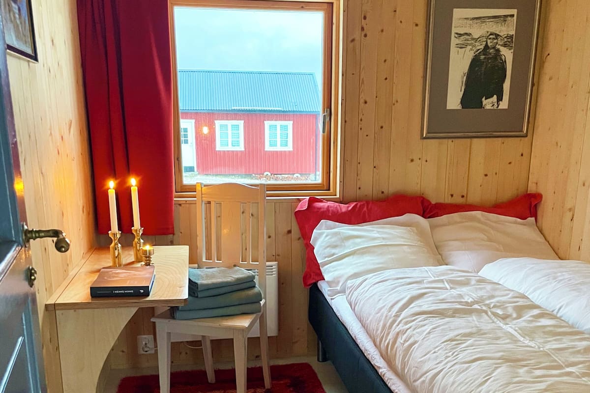 Una habitación pequeña y acogedora con una cama, un escritorio con silla, una ventana que ofrece vistas de un edificio rojo y un cuadro en la pared.