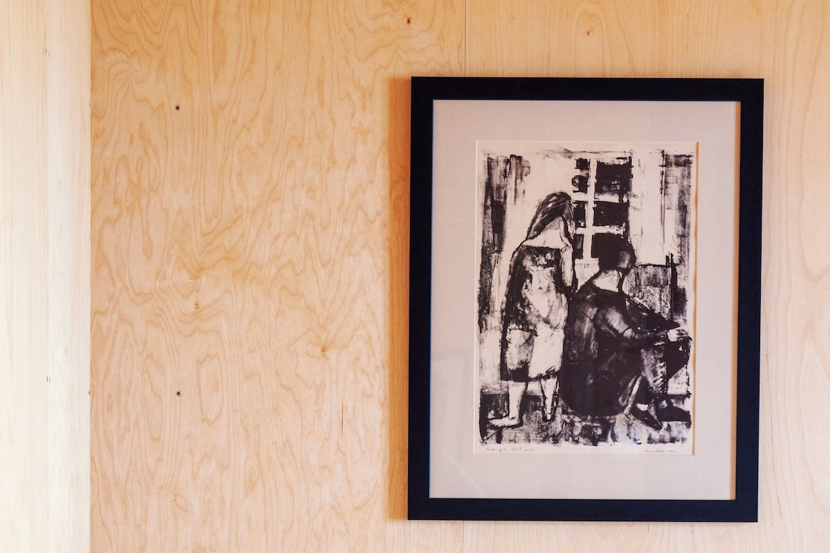 Pared de madera con una obra de arte blanco y negro enmarcada, una silla de madera con un jarrón de flores, y una cama con ropa blanca.