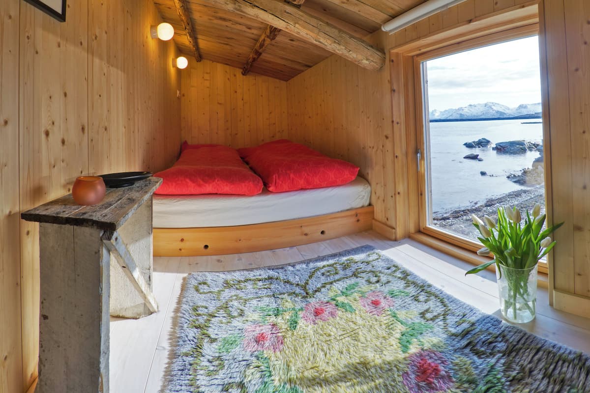 Habitación de una cabaña de madera con una cama, un tapete y una ventana con vistas a un cuerpo de agua y montañas.