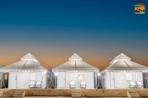 Three white tents with a peaked roof, each with two chairs and a small table, set against a twilight sky.