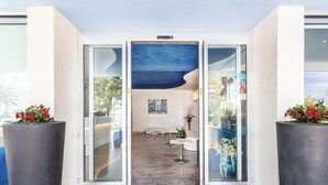 Un vestíbulo de hotel con diseño moderno, que presenta una entrada con puerta de vidrio, una mesa de recepción y una zona de estar con un cartel de 'welcome'.