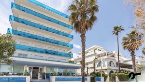 Ein modernes Hotel mit mehreren Balkonen, einem zentralen Eingang und Palmen.