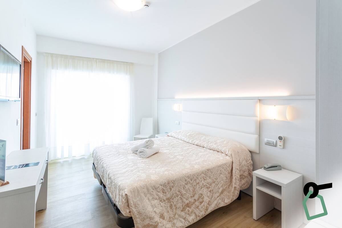 Una habitación de hotel moderna con una cama bien hecha, una mesita de noche con lámpara y un escritorio pequeño con una computadora portátil.