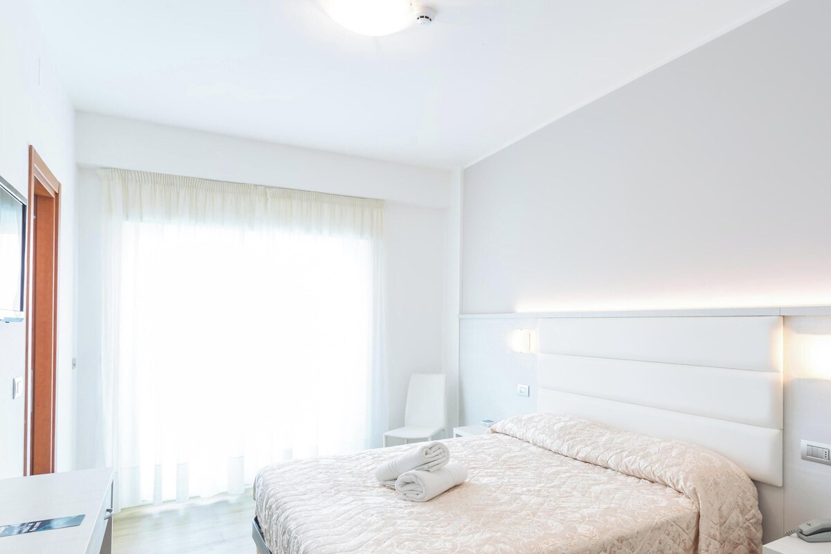 A modern hotel room with a neatly made bed, a bedside table with a lamp, and a small desk with a laptop.