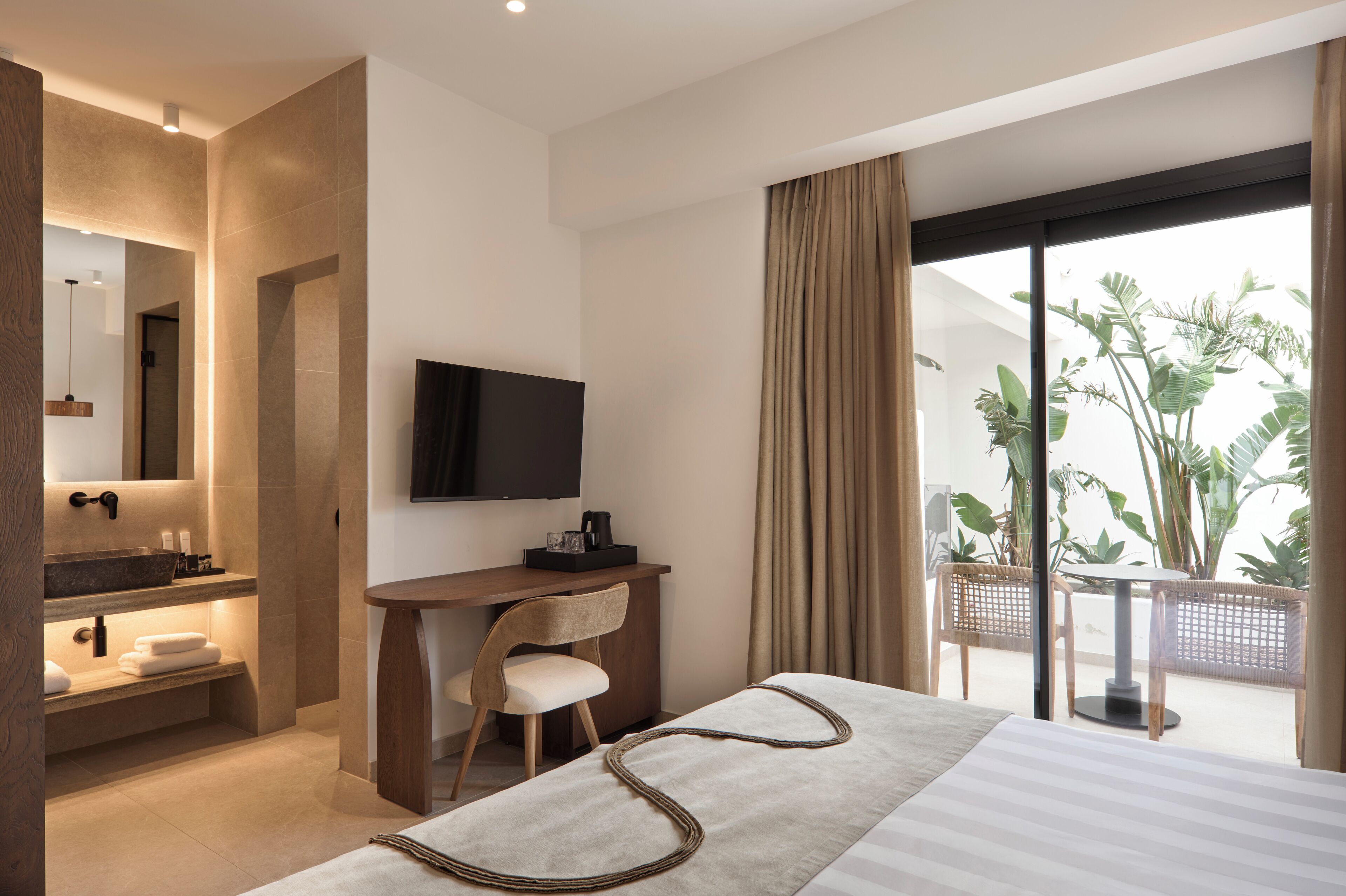 Een moderne hotelkamer met een groot bed, een bureau met een televisie, een badkamer met een wastafel en spiegel, en een balkon met planten.