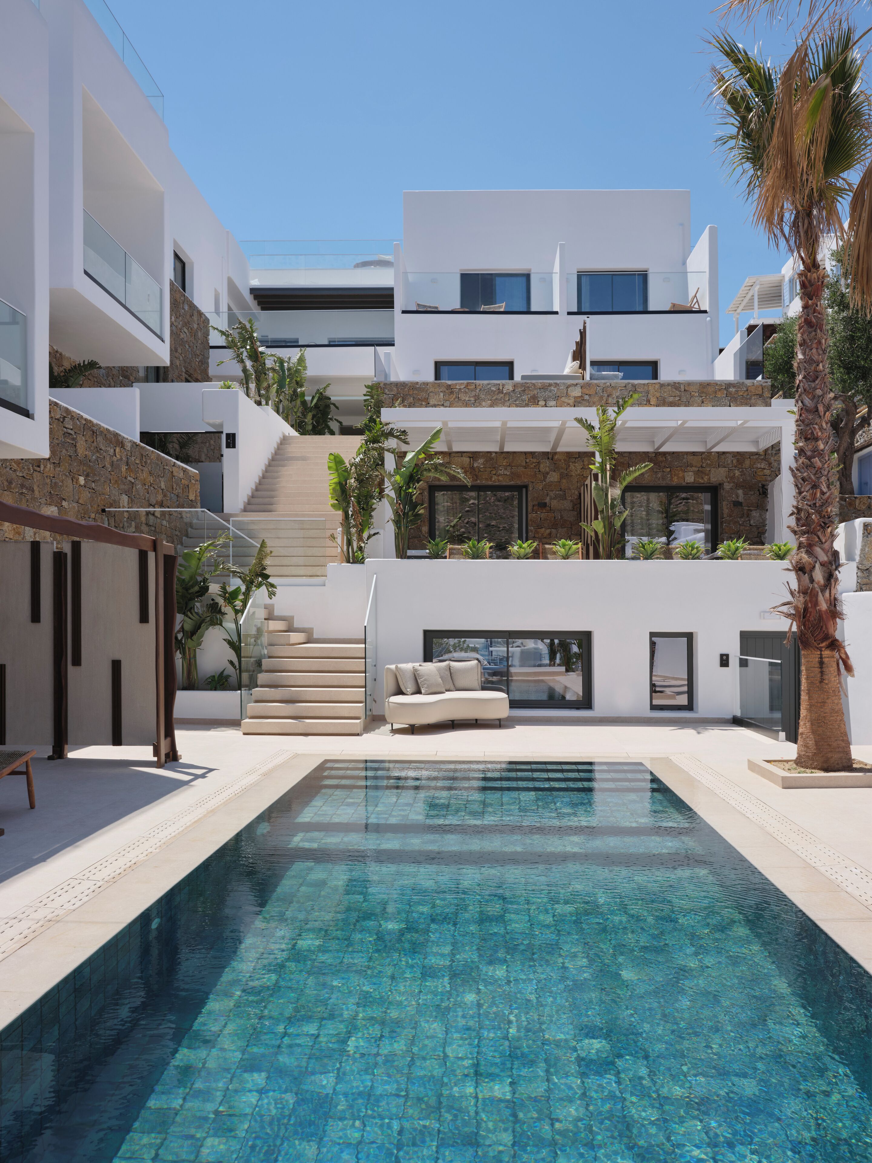 A modern pool area with a clear blue pool, a white outdoor seating area, and a staircase leading to a second-floor balcony.