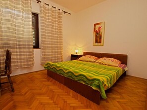 Un dormitorio con piso de madera en patrón de espiga, una cama con manta verde y amarilla a rayas, dos almohadas, una mesita de noche con lámpara, una silla y cortinas a rayas.