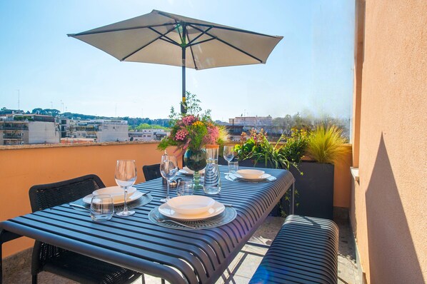 A rooftop terrace with a table set for dining, an umbrella, and potted plants.