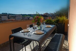 A rooftop terrace with a table set for two, a vase of flowers, and a view of the cityscape.