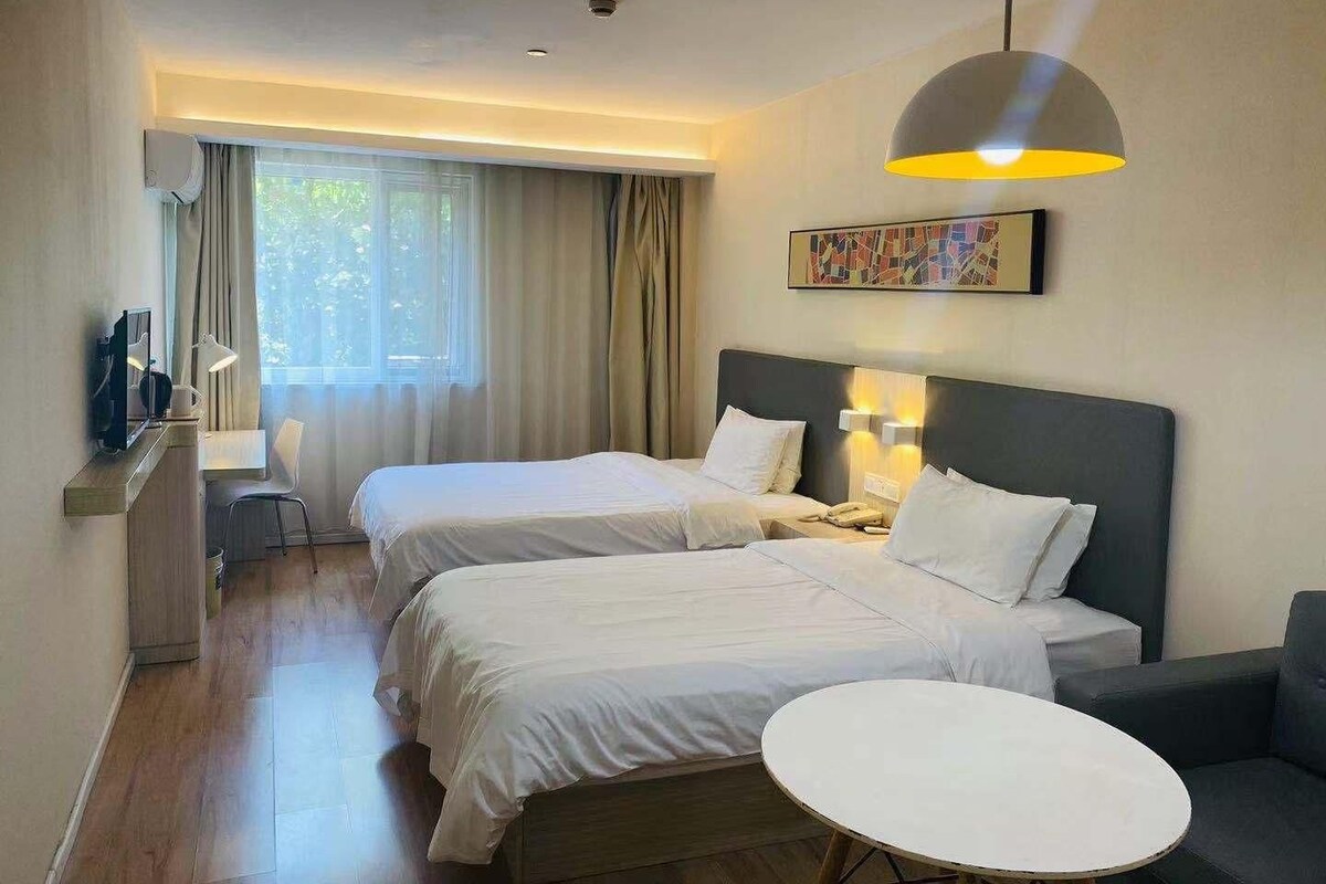 Ein Hotelzimmer mit zwei Betten, einem Schreibtisch mit Stuhl, einem kleinen runden Tisch und einem großen Fenster mit Vorhängen.
