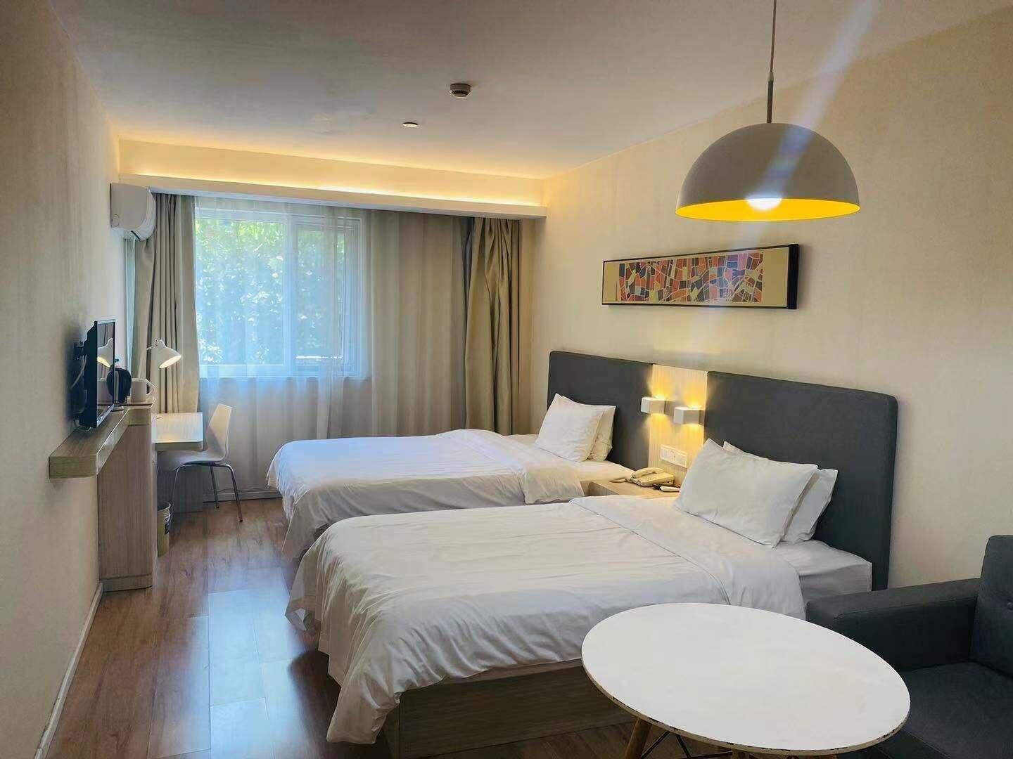 A hotel room with two beds, a desk with a chair, a small round table, and a large window with curtains.