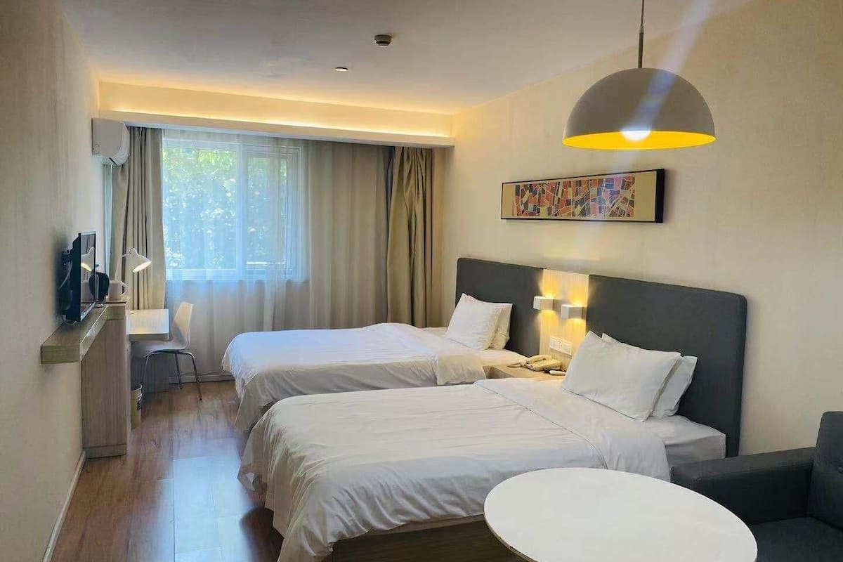 A hotel room with two beds, a desk with a chair, a small round table, and a large window with curtains.