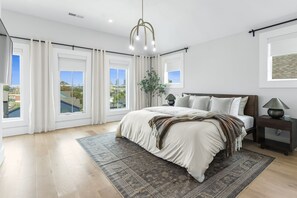 A bedroom with a large bed, a nightstand, a lamp, a potted plant, and large windows with curtains.