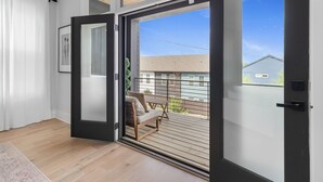 Une porte coulissante s’ouvrant sur un balcon doté d’un plancher en bois, d’une chaise et offrant une vue sur les maisons voisines.