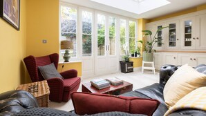 A living room with a sofa, armchair, and a coffee table. There is a window with a view of greenery outside. - Caithness Road VI by Onefinestay (London)