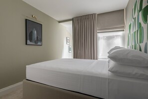 A neatly made bed with white linens in a room with a large window, beige curtains, and a framed picture on the wall. - Culford Gardens III by Onefinestay (London)
