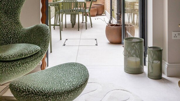 A modern interior with a green patterned chair, a table, and a lamp. There is a view of an outdoor area with a table and chairs, and a brick wall with greenery. - Culford Gardens III by Onefinestay (London)