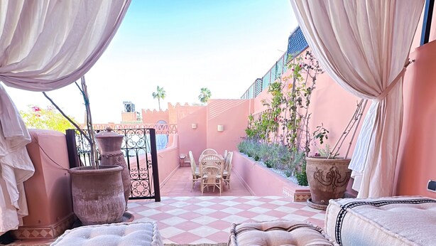A cozy outdoor seating area with a checkered floor, a cushioned bench, a wooden table with a tray of food and drinks, and a view of a garden and buildings in the background. - Riad Solila (Marrakech)