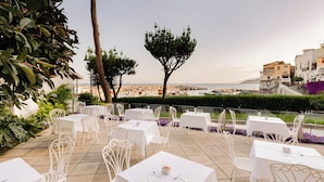 An outdoor seating area with white tables and chairs, overlooking a rocky coastline with buildings in the background. - Hotiday Room Collection - Massa Lubrense (Massa Lubrense)