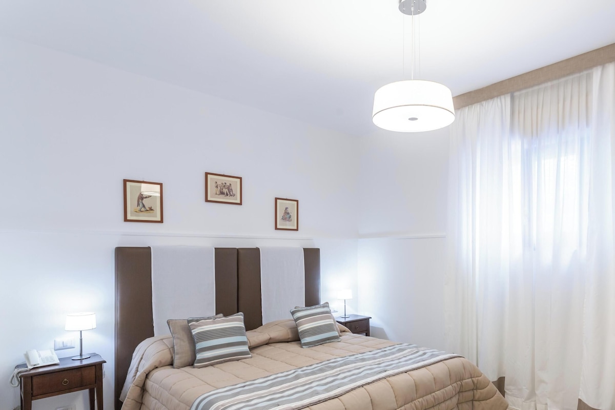 A hotel room with a large bed, a bedside table with a lamp, and a window with curtains.