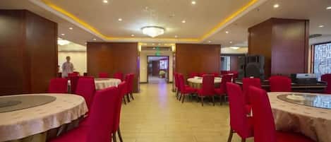 A hotel dining area with round tables and red chairs, a chandelier, and a tiled floor.
