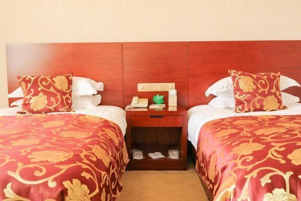A hotel room with two beds, each with a red and gold patterned bedspread, a wooden nightstand with a telephone and a green decorative item, and a wooden headboard.