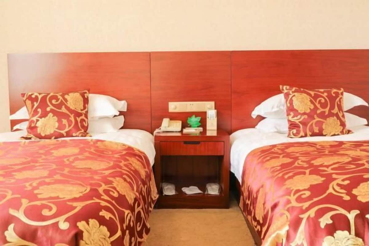 A hotel room with two beds, each with a red and gold patterned bedspread, a wooden nightstand with a telephone and a green decorative item, and a wooden headboard.