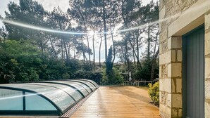 Una terraza de madera con cerramiento de vidrio, rodeada de árboles y un edificio de piedra.