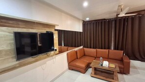 A modern living room with a flat-screen TV mounted on a wooden wall, a brown leather sofa, a wooden coffee table with a glass, and a ceiling fan. - Hotel Sapphire Inn (Gorakhpur)