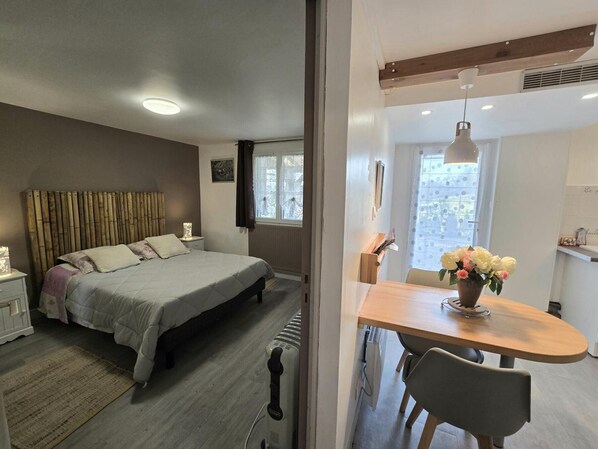 Un dormitorio con cama, mesita de noche y ventana con cortinas. Hay un comedor con mesa y sillas.