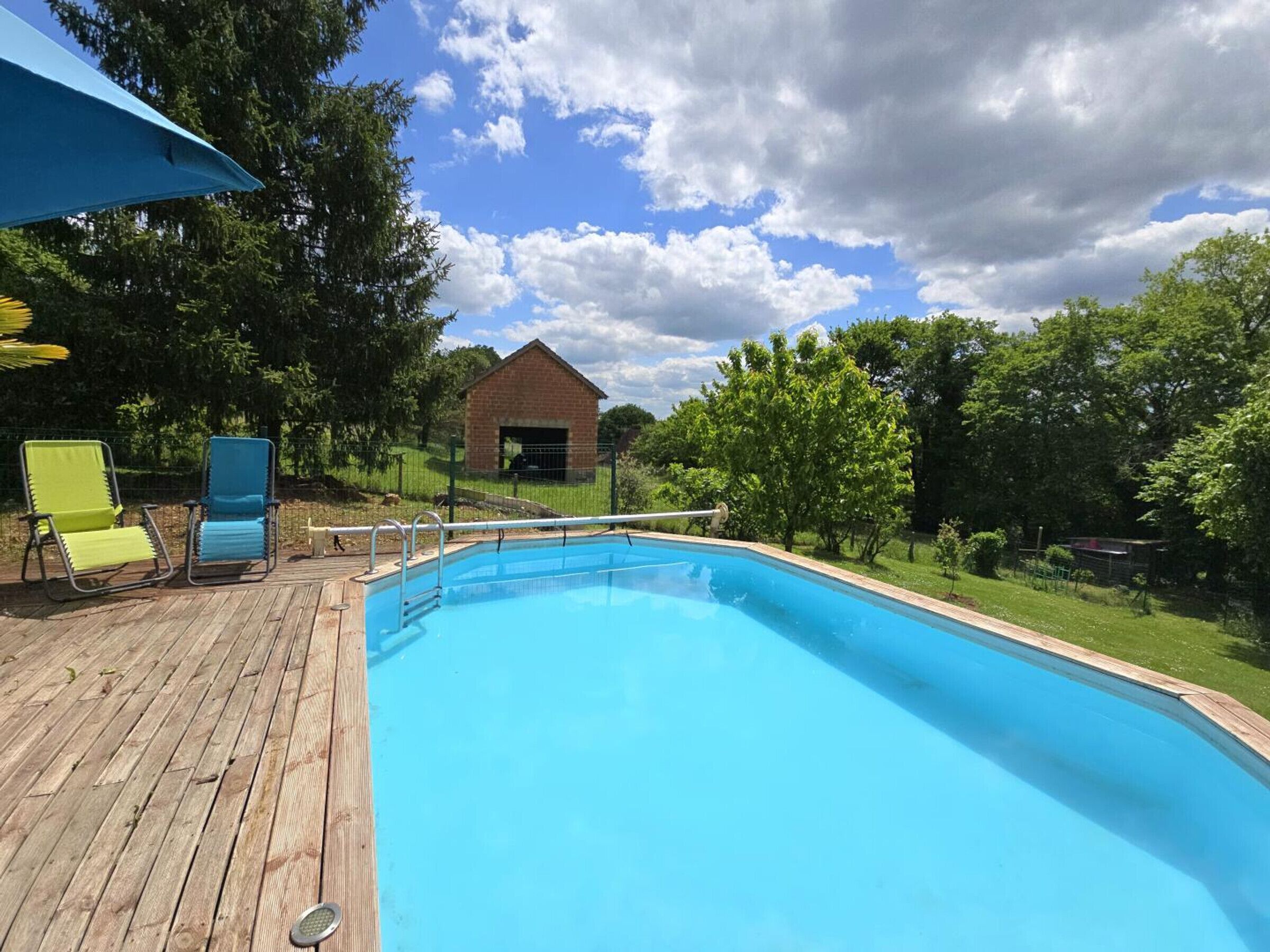 Una piscina con bordo in legno, due lettini da relax, un edificio in mattoni sullo sfondo e cielo azzurro con nuvole.
