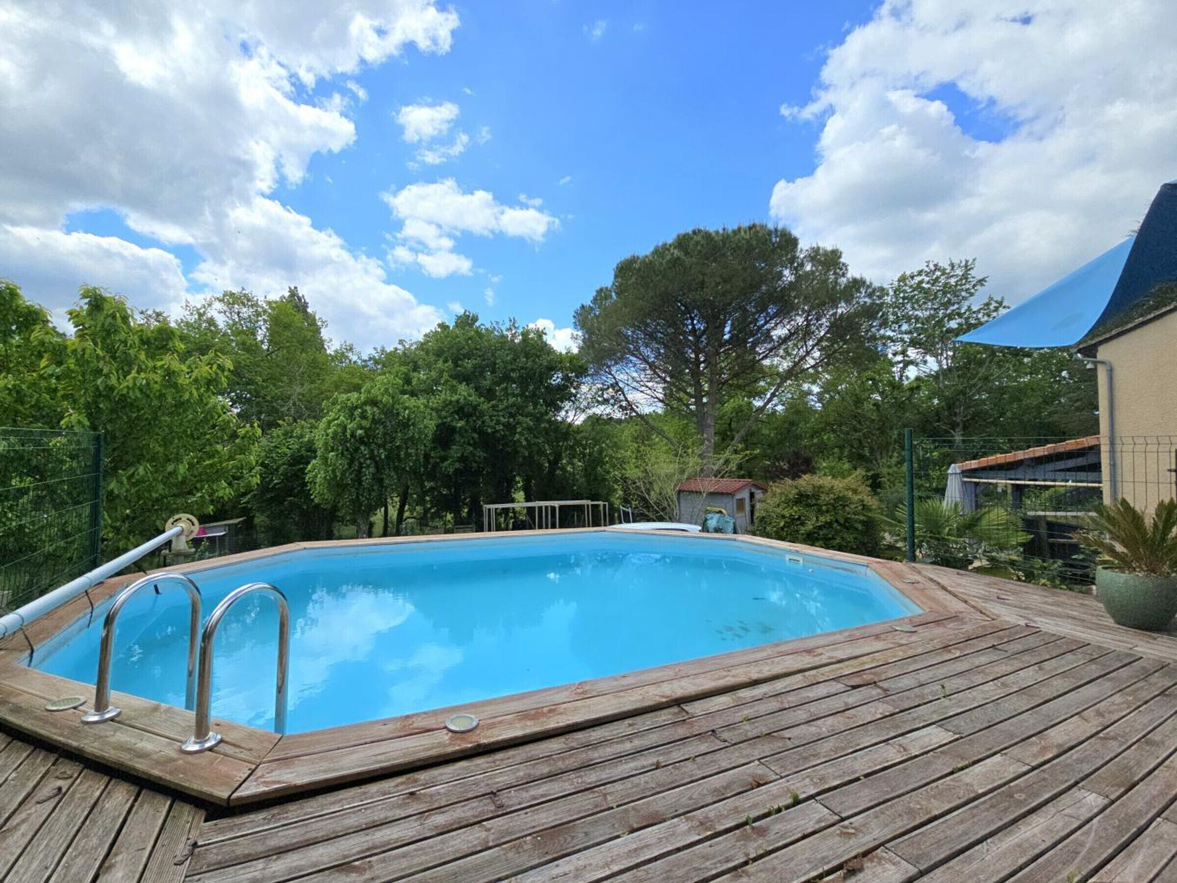 Une piscine dotée d’une terrasse en bois, entourée d’arbres et surplombée par un ciel dégagé.