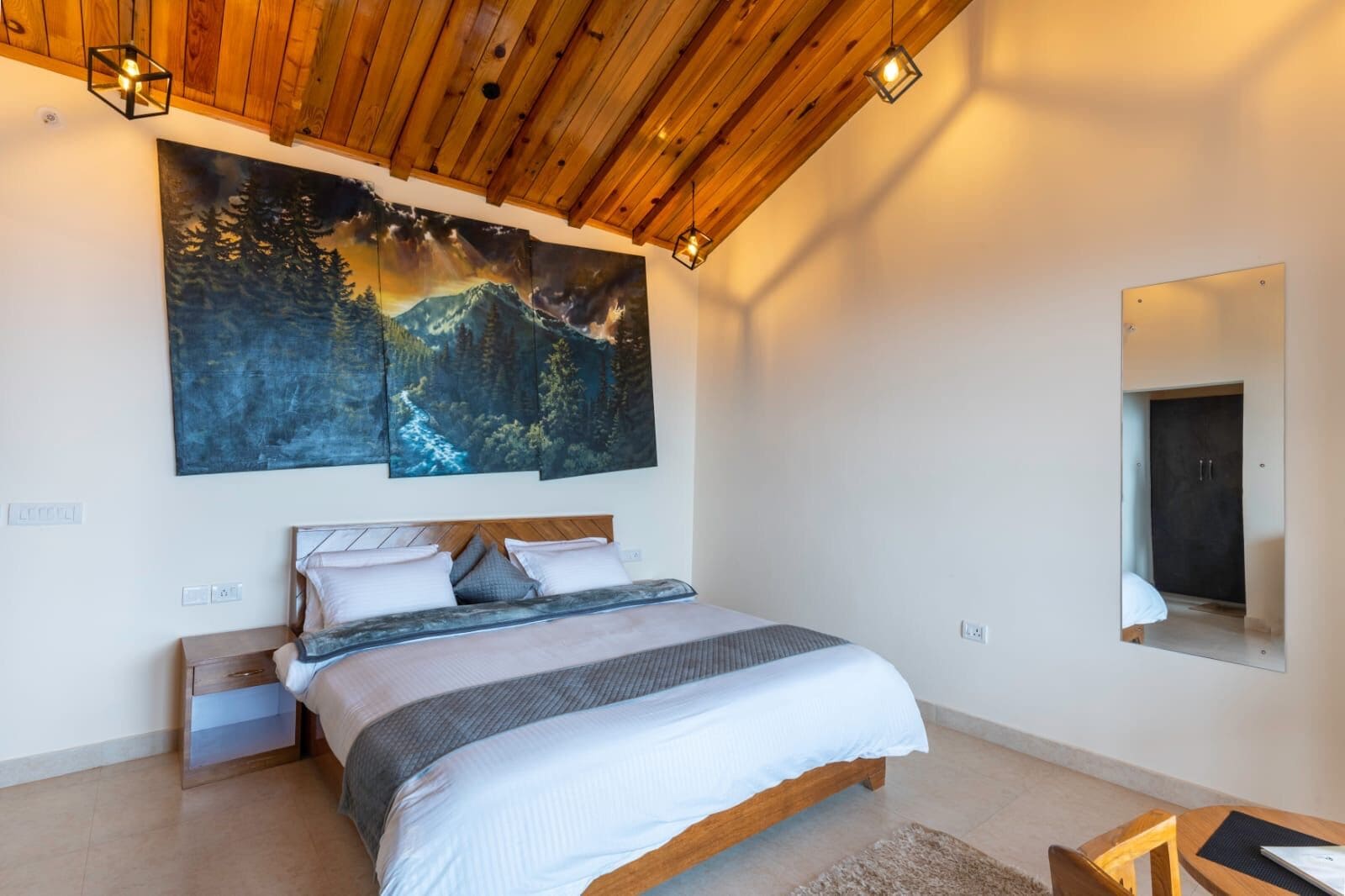 Un dormitorio con techo de madera, una cama grande con cabecera, una mesita de noche y una pintura de un paisaje forestal.