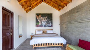 Un dormitorio con techo de madera, una cama con ropa de cama blanca, un cuadro de dos gatos en la pared, un cojín rojo y un banco de madera.