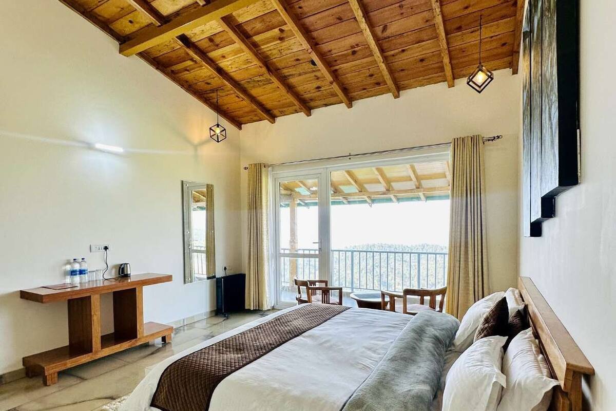 A spacious bedroom with a large bed, wooden ceiling beams, a wooden nightstand, and a view of the outdoors through large windows.