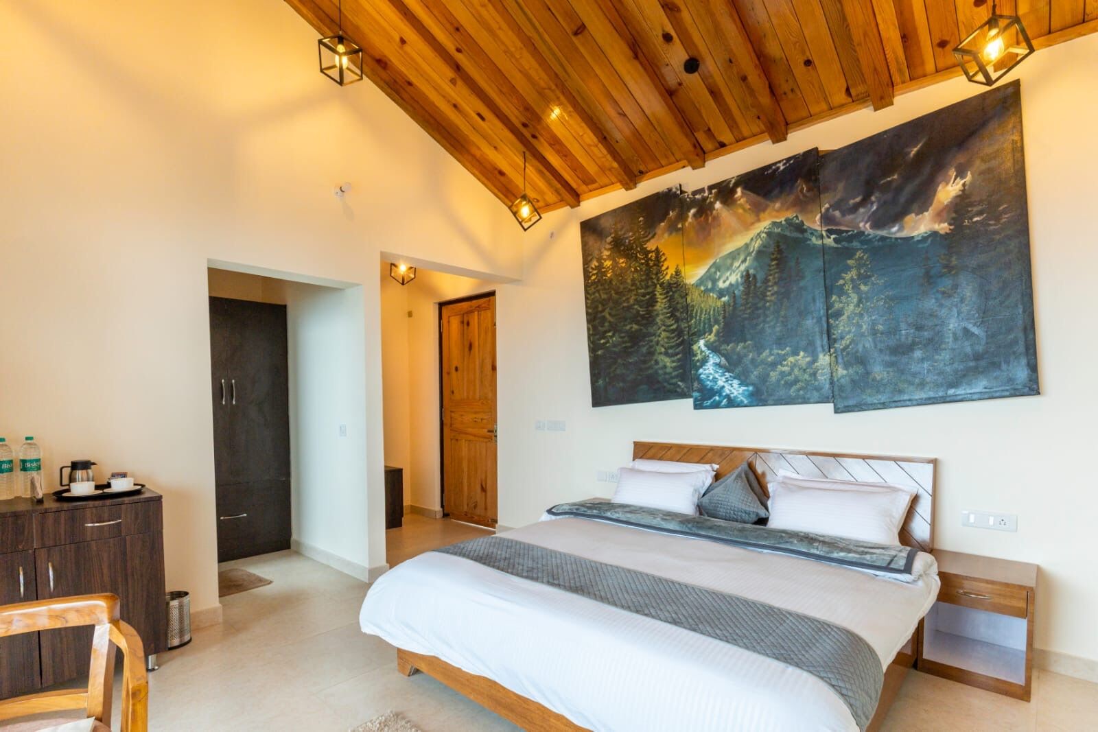 A bedroom with a wooden ceiling, a large bed with pillows, a nightstand, a dresser, and a painting of a forest scene.