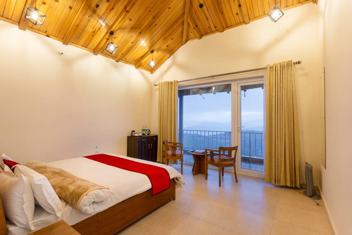 A bedroom with a wooden ceiling, a bed with a red blanket, a small table with chairs, and a view of the sea through a glass door.