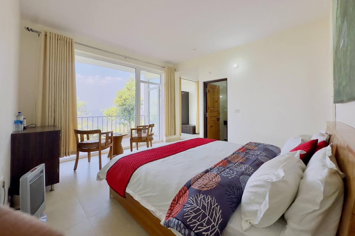 A hotel room with a large bed, a desk, a chair, and a balcony with a view of trees.