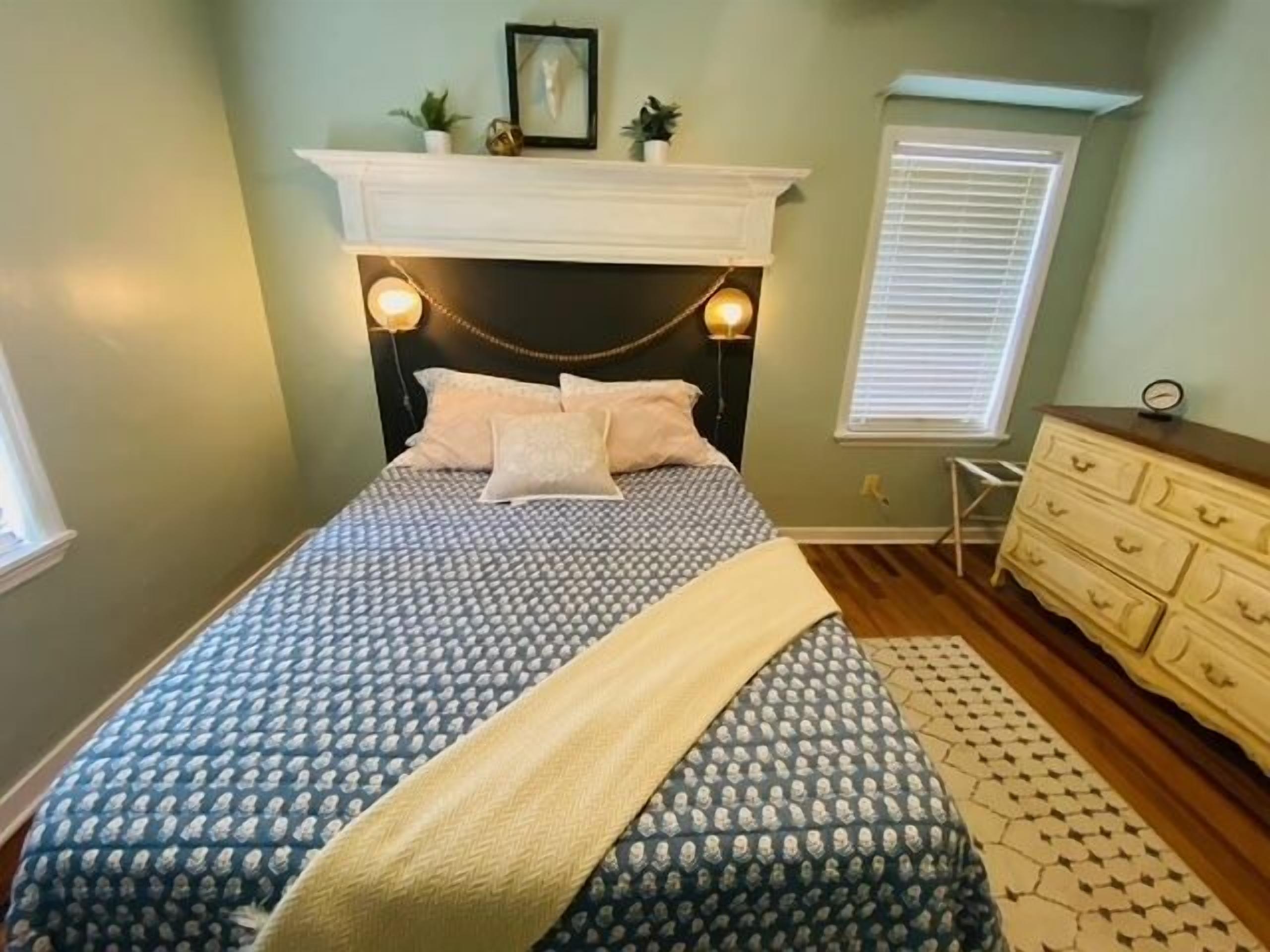 A bedroom with a patterned bedspread, a headboard with wall-mounted lights, a dresser, and a window with blinds.